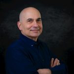 Bald man wearing a blue shirt, smiling confidently with crossed arms in a studio setting.
